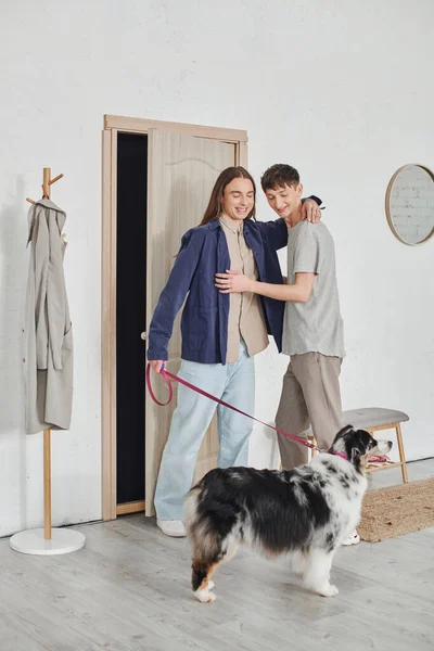 Glückliches schwules Paar in lässigem Outfit, das im Flur neben der Garderobe steht und sich umarmt und in der Nähe eines australischen Schäferhundes an der Leine hält und in einer modernen Wohnung zusammen lächelt — Stockfoto