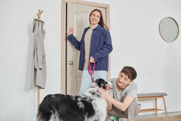 Feliz gay homem sorrindo enquanto abraçando adorável australiano pastor cão ao lado de alegre namorado com longo cabelo segurando trela enquanto de pé perto da porta no moderno corredor — Fotografia de Stock