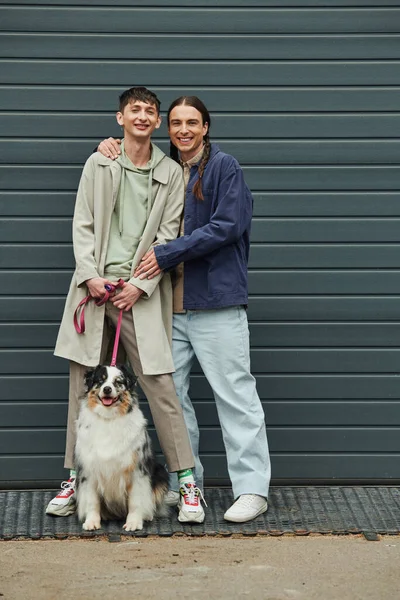 Cheerful gay man with pigtails hugging smiling tattooed boyfriend in casual outfit holding leash of Australian shepherd dog and standing next to near garage door outside on street — Stock Photo