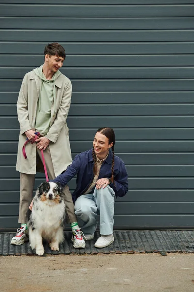 Gai gay l'homme avec des nattes câlins australien berger chien à côté de sourire copain dans manteau tenant laisse près de garage porte à l'extérieur sur rue — Photo de stock