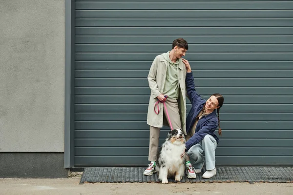 Happy and young gay man with pigtails cuddling Australian shepherd dog next to smiling boyfriend in coat holding leash near garage door outside on street — Stock Photo