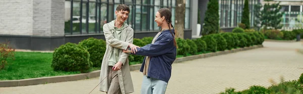 Happy lgbt couple holding hands of each other while walking out together and smiling near green bushes and modern building on blurred background on urban street, banner — Stock Photo