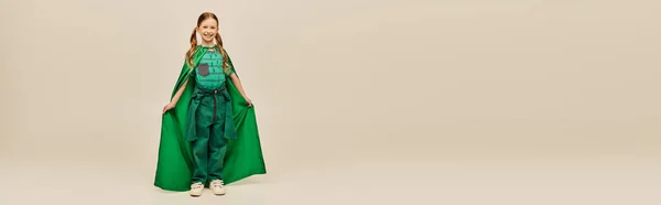 Menina sorridente em traje de super-herói verde com manto vestindo calças e camiseta e de pé enquanto celebra o feriado do dia de proteção da criança em fundo cinza, banner — Fotografia de Stock