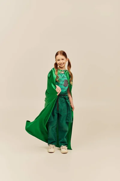 Glückliches Kind im grünen Superheldenkostüm mit Mantel, Hose und T-Shirt und stehend beim Kinderschutztag auf grauem Hintergrund — Stockfoto