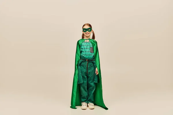 Menina sorridente em traje de super-herói verde com capa e máscara no rosto, vestindo calças e camiseta e de pé enquanto celebra o feriado do dia de proteção da criança no fundo cinza — Fotografia de Stock