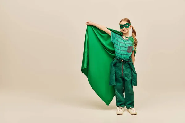 Happy kid in superhero costume and mask on face holding green cloak, wearing pants and t-shirt and standing while celebrating Child protection day holiday on grey background — Stock Photo