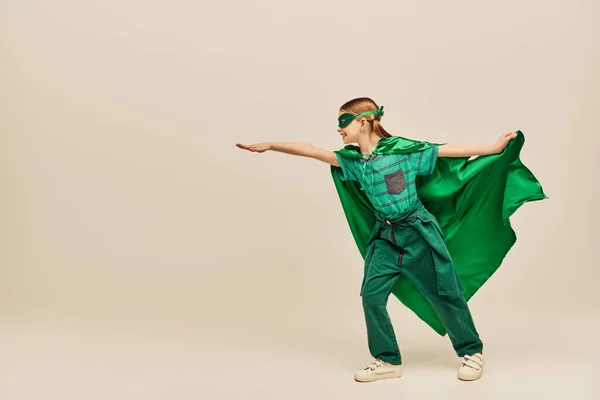 Вид збоку щасливої дитини в костюмі супергероя і маска, що тримає зелений плащ і стоїть з простягнутою рукою, святкуючи день захисту дитини на сірому фоні — стокове фото