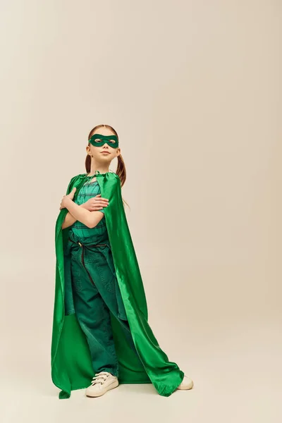 Серьезная девушка в зеленом костюме супергероя с плащом и маской на лице, стоящей со сложенными руками и смотрящей в камеру во время празднования Дня защиты детей на сером фоне — стоковое фото