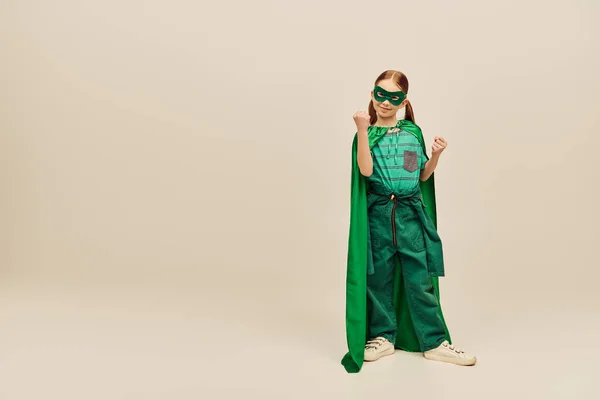 Powerful girl in green superhero costume with cloak and mask on face, wearing pants and t-shirt and standing with clenched fists while celebrating Child protection day holiday on grey background — Stock Photo
