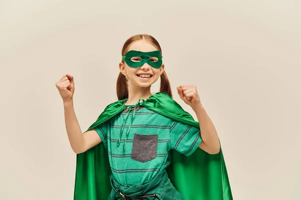 Poderosa chica en traje de superhéroe verde con capa y máscara en la cara, sonriendo y de pie con los puños cerrados mientras se celebra la fiesta del Día Mundial de la Protección del Niño sobre fondo gris - foto de stock