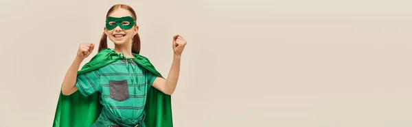 Garoto poderoso em traje de super-herói verde com capa e máscara no rosto, sorrindo e de pé com punhos apertados enquanto celebra as férias do Dia Mundial da Proteção da Criança no fundo cinza, banner — Fotografia de Stock