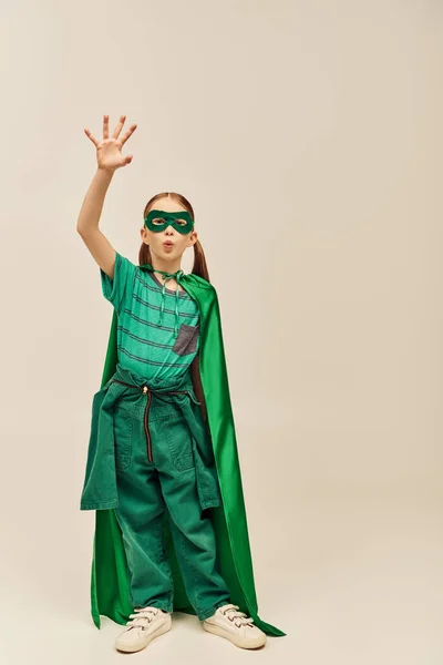 Здивована дівчина в зеленому костюмі супергероя з плащем і маскою на обличчі, кидає губи і стоїть з витягнутою рукою під час святкування дня захисту дітей на сірому фоні — Stock Photo