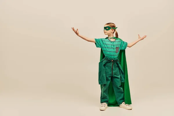 Сильная девушка в костюме супергероя с зеленым плащом и маской на лице, стоящая с протянутыми руками, демонстрируя силу и отмечая Международный день защиты детей на сером фоне — стоковое фото