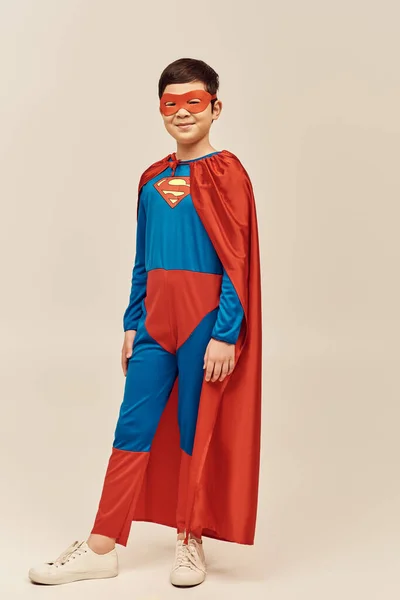 In voller Länge glücklicher asiatischer Junge im rot-blauen Superheldenkostüm mit Mantel und Maske auf dem Gesicht, der lächelnd den Internationalen Kindertag auf grauem Hintergrund feiert — Stockfoto