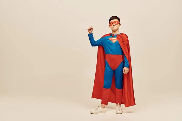 Mächtiger asiatischer Junge im rot-blauen Superheldenkostüm mit Mantel und Maske auf dem Gesicht, der eine starke Geste zeigt, während er den Internationalen Kindertag auf grauem Hintergrund feiert — Stockfoto