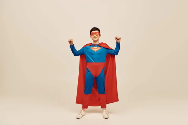Glücklicher asiatischer Junge in rot-blauem Superheldenkostüm mit Mantel und Maske auf dem Gesicht, der eine Kraftgeste zeigt, während er Happy Children 's Day auf grauem Hintergrund feiert — Stockfoto