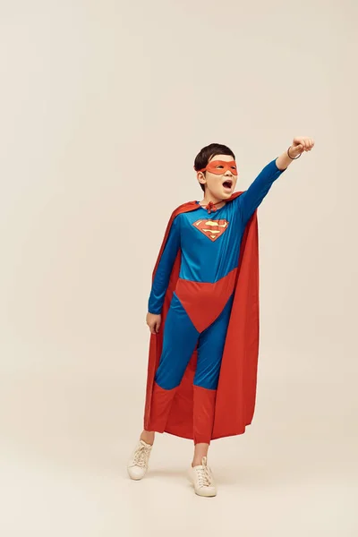 Menino asiático em traje de super-herói com capa e máscara no rosto gritando enquanto mostra gesto de força enquanto está de pé com a mão estendida no fundo cinza, conceito de dia de proteção infantil — Fotografia de Stock