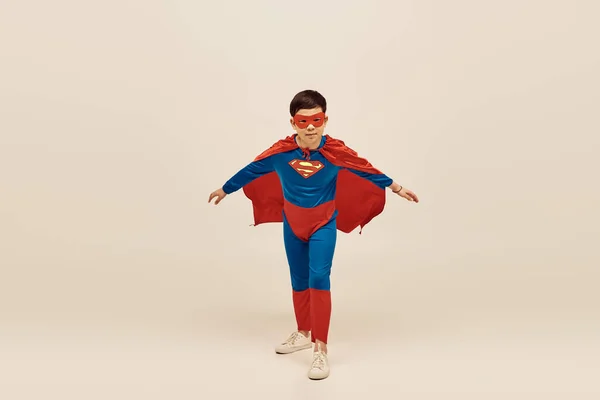 Мужественный азиатский мальчик в красно-синем супергеройском костюме с плащом и маской на лице, отмечающий Международный день защиты детей на сером фоне — стоковое фото