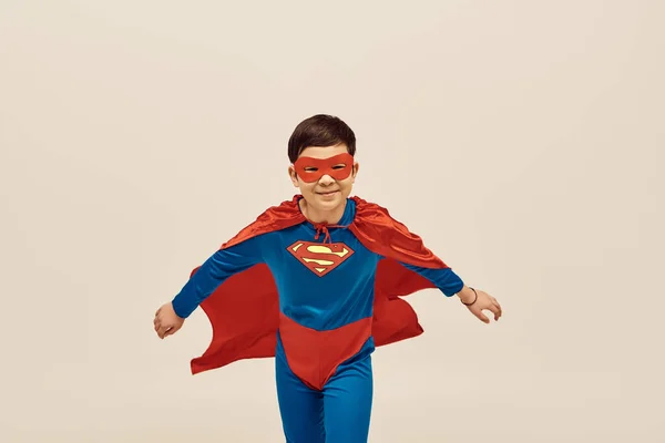 Niño asiático feliz en traje de superhéroe rojo y azul con capa y máscara en la cara sonriendo mientras se celebra el Día Internacional de la Protección de los Niños sobre fondo gris - foto de stock