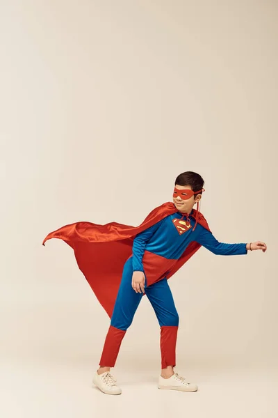 In voller Länge glücklicher asiatischer Junge im Superheldenkostüm mit Mantel und Maske lächelt beim Wegschauen und steht gegen den Wind während des Internationalen Kinderferientages auf grauem Hintergrund — Stockfoto