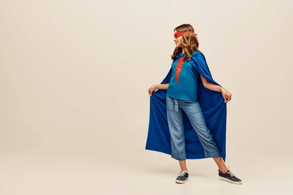 Menina feliz em traje de super-herói e máscara vermelha no rosto segurando capa azul, de pé em jeans jeans jeans e t-shirt no fundo cinza, o conceito do Dia Internacional para a Proteção das Crianças — Fotografia de Stock
