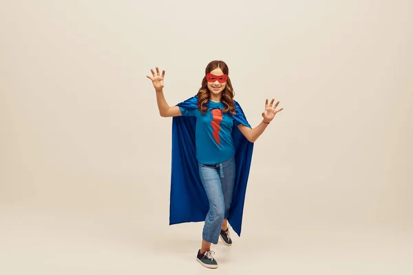 Fröhliches Mädchen im Superheldenkostüm mit blauem Mantel und roter Maske im Gesicht, mit Handbewegung, stehend in Jeans und T-Shirt beim Kinderschutztag auf grauem Hintergrund — Stockfoto