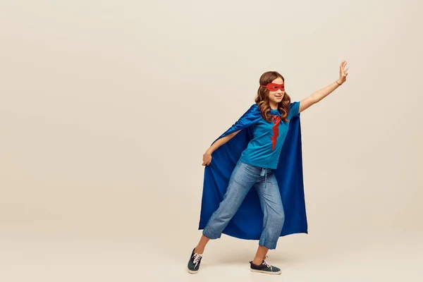 Chica feliz en traje de superhéroe con capa azul y máscara roja en la cara, de pie en vaqueros y camiseta con la mano extendida, mientras que llegar a algo en fondo gris - foto de stock