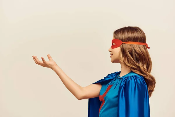 Вид сбоку счастливой девушки в костюме супергероя с синим плащом и красной маской на лице, стоящей с протянутой рукой во время на сером фоне в студии, концепция Всемирного дня защиты детей — стоковое фото