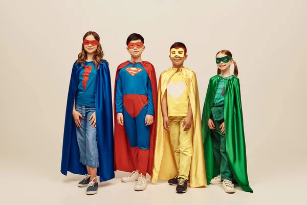 Heureux interracial préadolescents enfants en costumes de super-héros colorés avec des manteaux et des masques debout ensemble tout en célébrant la Journée de la protection de l'enfance vacances sur fond gris en studio — Photo de stock
