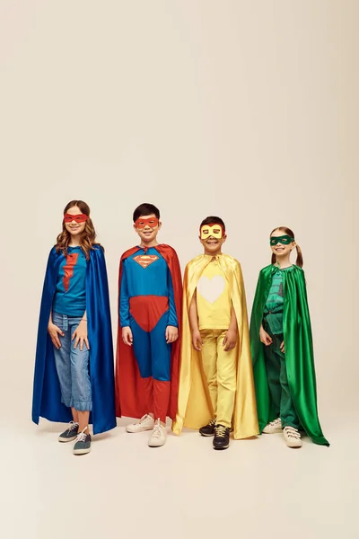Niños preadolescentes multiétnicos felices en trajes coloridos de superhéroes con capas y máscaras de pie juntos mientras celebran las vacaciones del día de protección infantil sobre fondo gris en el estudio - foto de stock