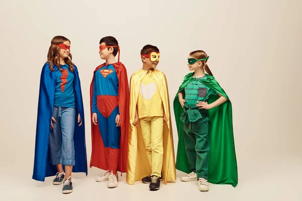 Crianças pré-adolescentes inter-raciais felizes em trajes coloridos de super-heróis com capas e máscaras de pé e olhando uns para os outros em fundo cinza no estúdio, Dia Mundial da Proteção à Criança — Fotografia de Stock