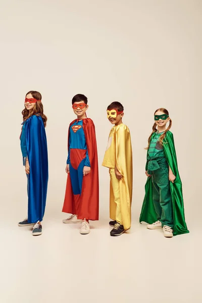 Веселі міжрасові діти в барвистих костюмах супергероїв з плащами і масками, що стоять разом і дивляться на камеру, святкуючи день захисту дітей на сірому фоні в студії — стокове фото