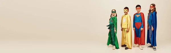 Feliz interracial pré-adolescente crianças em trajes coloridos com capas e máscaras de pé juntos enquanto celebra o feriado do dia de proteção da criança em fundo cinza no estúdio, banner — Fotografia de Stock