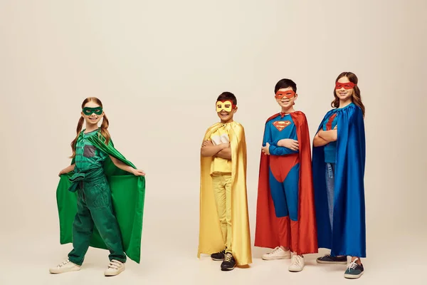 Счастливая девушка, стоящая в зеленом плаще и маске рядом с межрасовыми друзьями-подростками в красочных костюмах супергероев во время празднования Дня защиты детей на сером фоне в студии — стоковое фото