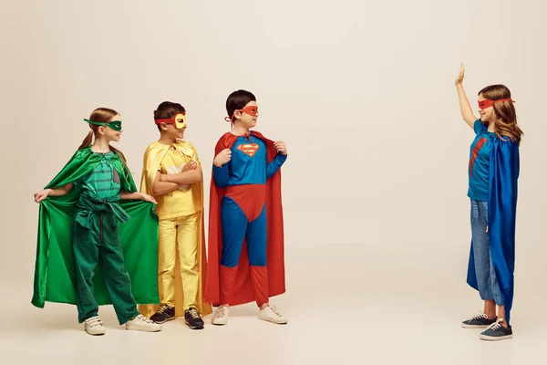 Crianças pré-adolescentes inter-raciais felizes em trajes coloridos de super-heróis com capas e máscaras olhando para a menina acenando mão no fundo cinza no estúdio, o conceito do Dia Internacional para a Proteção das Crianças — Fotografia de Stock