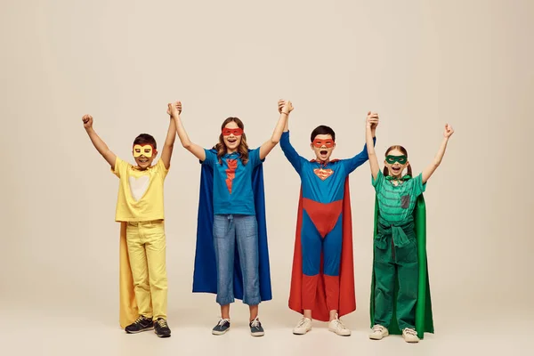 Счастливые межрасовые дети в красочных костюмах супергероев в плащах и масках, держащиеся за руки и смотрящие в камеру на сером фоне в студии, концепция Международного дня защиты детей — стоковое фото