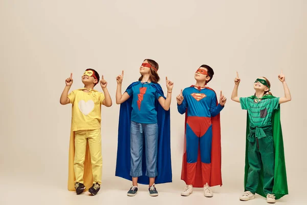 Positive multicultural kids in colorful superhero costumes with cloaks and masks pointing with fingers while celebrating Child protection day holiday on grey background in studio — Stock Photo