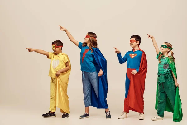 Positive multicultural kids in colorful superhero costumes with cloaks and masks pointing with fingers while looking away on grey background in studio, International Day for Protection of Children — Stock Photo