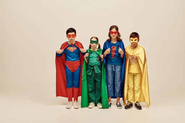 Bambini multiculturali in costumi colorati con mantelli e maschere che fanno male alle labbra, guardano insieme la macchina fotografica e celebrano la giornata internazionale dei bambini su sfondo grigio in studio — Foto stock