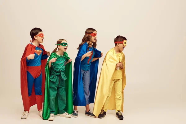 Valientes niños multiculturales en trajes coloridos con capas y máscaras gritando y de pie con los puños apretados juntos sobre fondo gris en el estudio, concepto del Día de la Protección del Niño - foto de stock