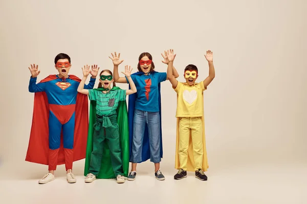 Мужественные мультикультурные дети в красочных костюмах в плащах и масках, кричащие и поднимающие руки вместе на сером фоне в студии, концепция Дня защиты детей — стоковое фото