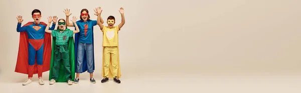Mutige Kinder in bunten Kostümen mit Mänteln und Masken, die vor grauem Hintergrund im Atelier schreien und die Hände heben, Konzept Kinderschutztag, Transparent — Stockfoto