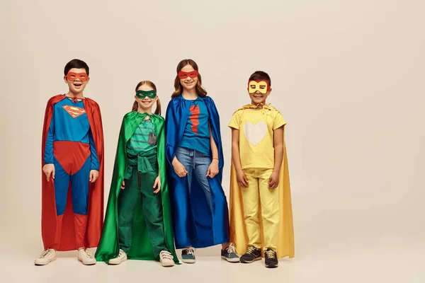 Happy interracial kids in colorful costumes with cloaks and masks smiling together and looking at camera on grey background in studio, Child Protection Day concept — Stock Photo