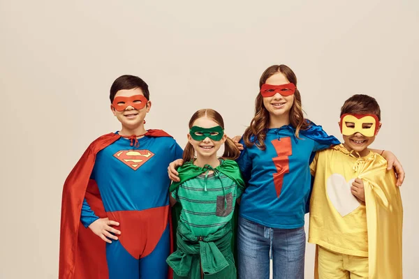 Happy interracial kids in colorful costumes with cloaks and masks smiling and hugging each other while looking at camera on grey background in studio, Child Protection Day concept — Stock Photo