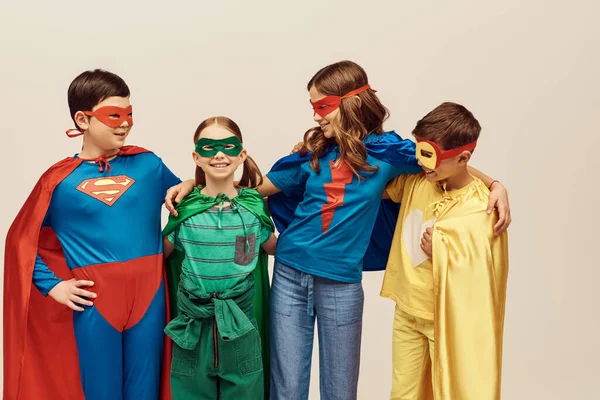 Crianças inter-raciais felizes em trajes coloridos com capas e máscaras sorrindo e abraçando menina alegre em fundo cinza no estúdio, conceito de Dia de Proteção à Criança — Fotografia de Stock