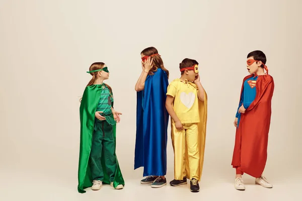 Chocado interracial crianças no colorido super-herói trajes com capas e máscaras olhando para o outro no fundo cinza no estúdio, conceito de dia internacional das crianças — Fotografia de Stock