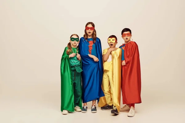 Shocked multicultural and preteen kids in colorful superhero costumes with cloaks and masks looking at camera on grey background in studio, International children's day concept — Stock Photo