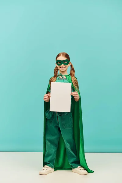 Щаслива дівчина в зеленому костюмі супергероя з плащем і маскою, що стоїть з чистим папером і дивиться на камеру, святкуючи день захисту дітей на синьому фоні — Stock Photo