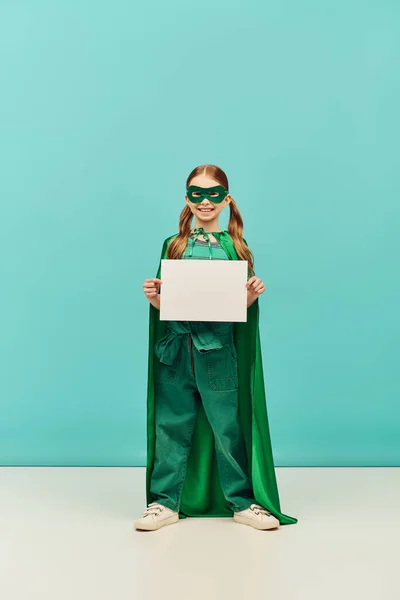 Joyeuse fille en costume de super-héros vert avec manteau et masque debout avec du papier vierge et regardant la caméra tout en célébrant les vacances de la journée de protection de l'enfance sur fond bleu — Photo de stock