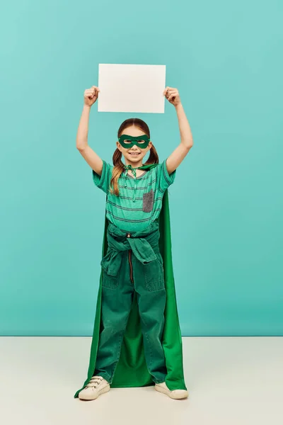 Happy preteen girl in green superhero costume with cloak and mask holding blank paper above head and looking at camera while celebrating Child protection day holiday on blue background — Stock Photo